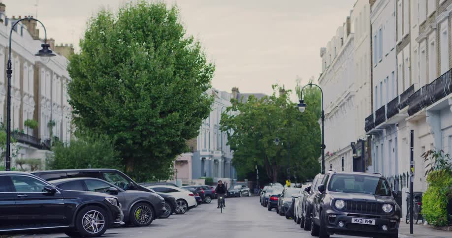 A young man rides his bicycle down a quiet, tree-lined residential street in London, flanked by parked cars and classic terraced houses