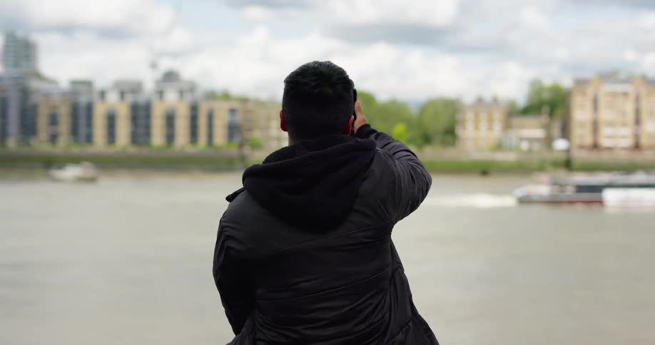 A man sits with his back to the camera, taking a picture with his smartphone of a boat passing by on a river in front of a city skyline