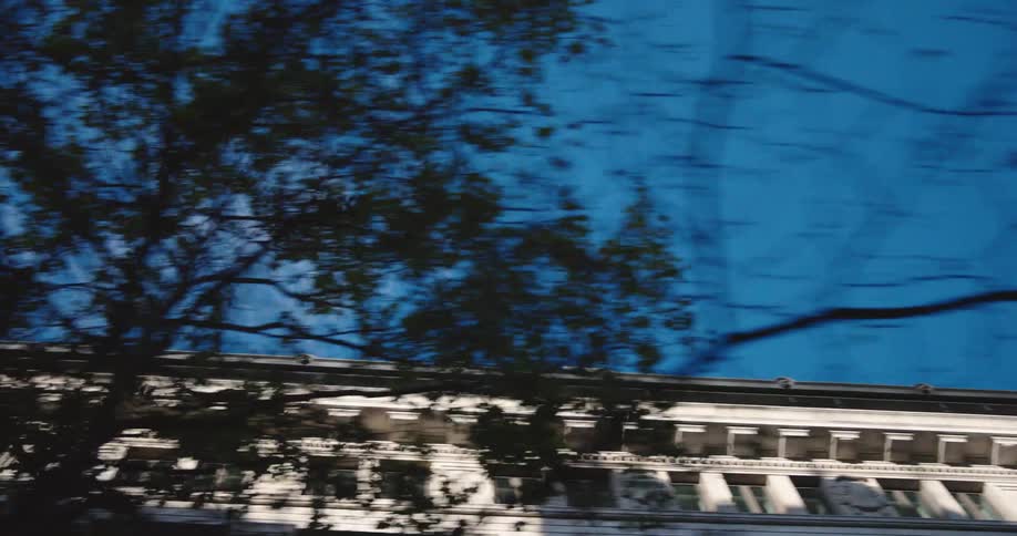 A slow motion tracking shot looks up at the top of a classic city building against a clear blue sky as trees pass by
