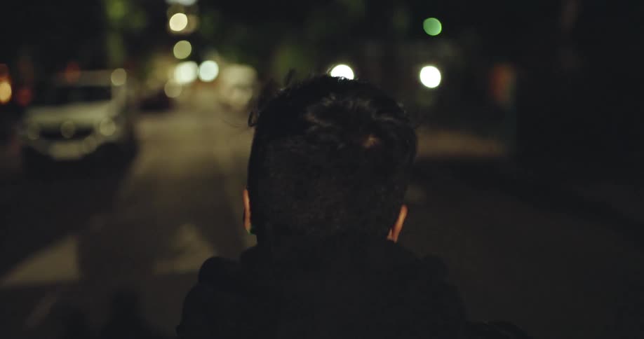 Slow motion over-the-shoulder shot of a young man walking down a dimly lit city street at night
