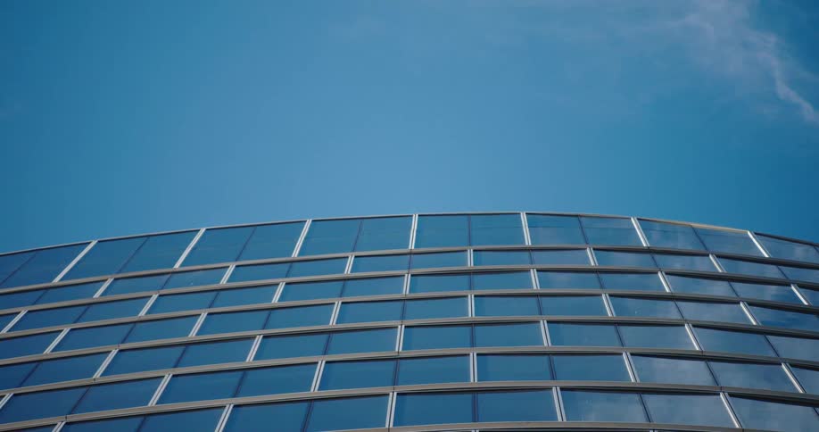 A low-angle cinemagraph shows wispy clouds drifting across a clear blue sky above a modern curved glass building