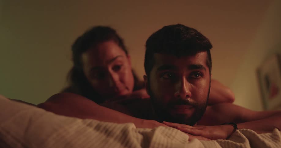 A thoughtful man lies in bed with his partner behind him, engaged in a serious conversation in a dimly lit room