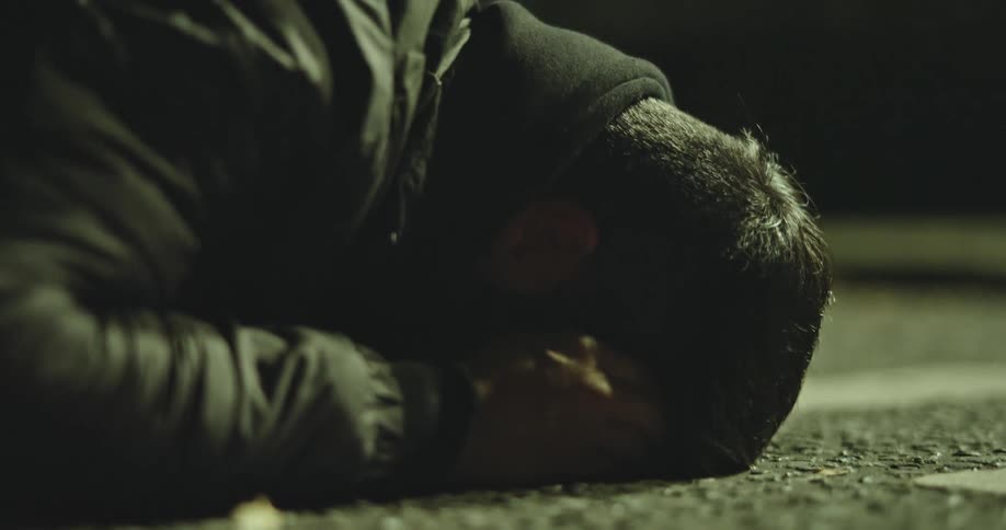 A man in a dark hoodie lies in despair on the pavement at night, slowly lifting his head
