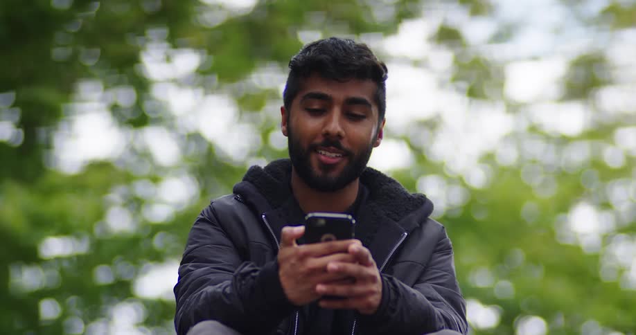 A happy young man in a black jacket uses his smartphone, smiling and texting before taking a selfie in a park