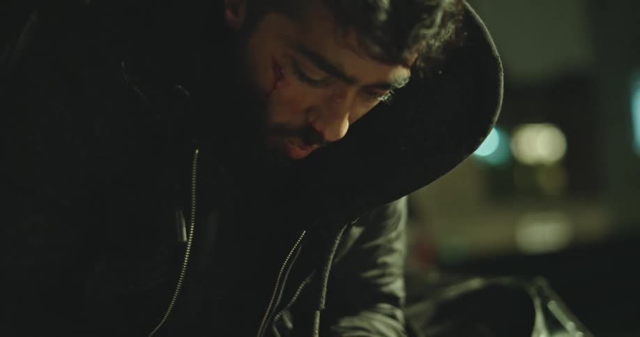 An injured and hungry man in a hoodie desperately eats a sandwich in slow motion at night