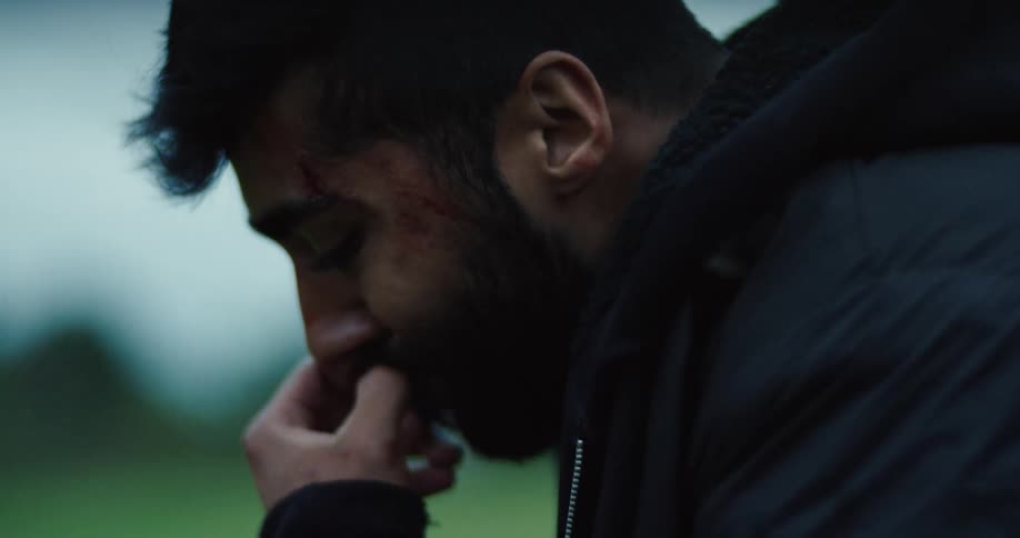 A close-up profile of a young man with a wounded face, looking down in sadness and contemplation on an overcast day