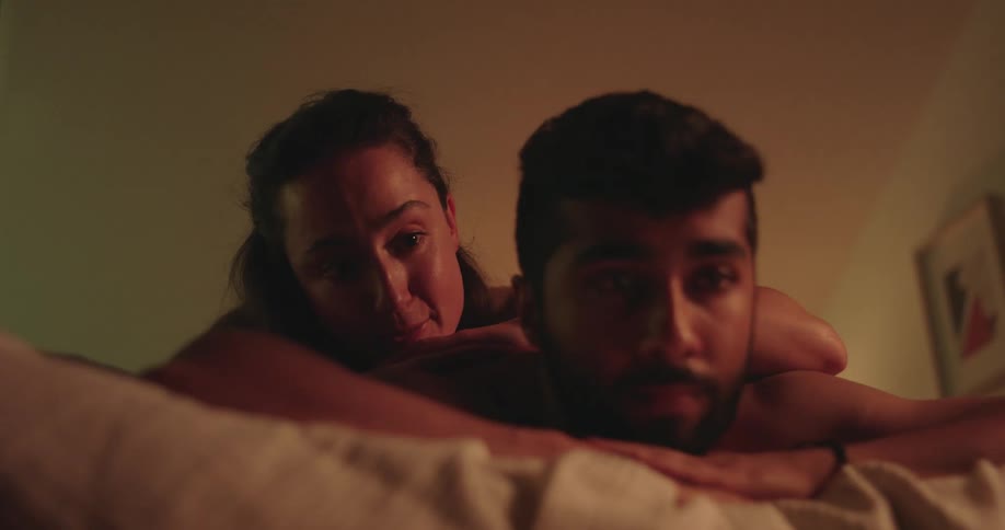 An intimate and somber close-up of a woman comforting her pensive partner as they lie together in a softly lit bedroom at night