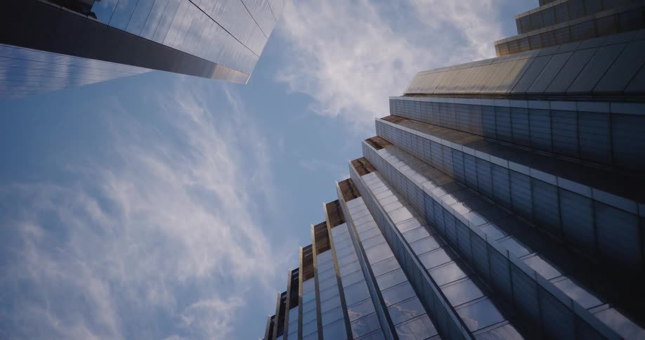A low-angle shot looks up at two towering glass skyscrapers reflecting a blue sky with wispy clouds passing by