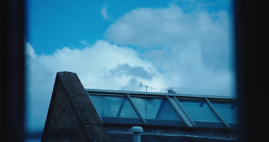 A crescent moon is visible in the cloudy blue daytime sky, as seen from a window looking out over a rooftop with skylights