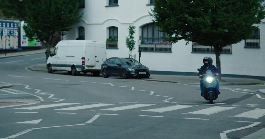 Two men in helmets ride a black electric scooter through a city street in slow motion