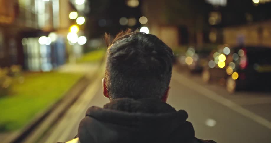Over-the-shoulder shot of a man in a hoodie walking down a street at night with city lights in the background