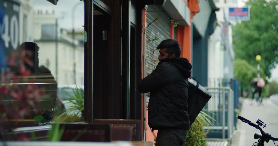 A male food delivery courier picks up a takeout order from a restaurant, places it in his insulated backpack, and prepares to leave on his bicycle