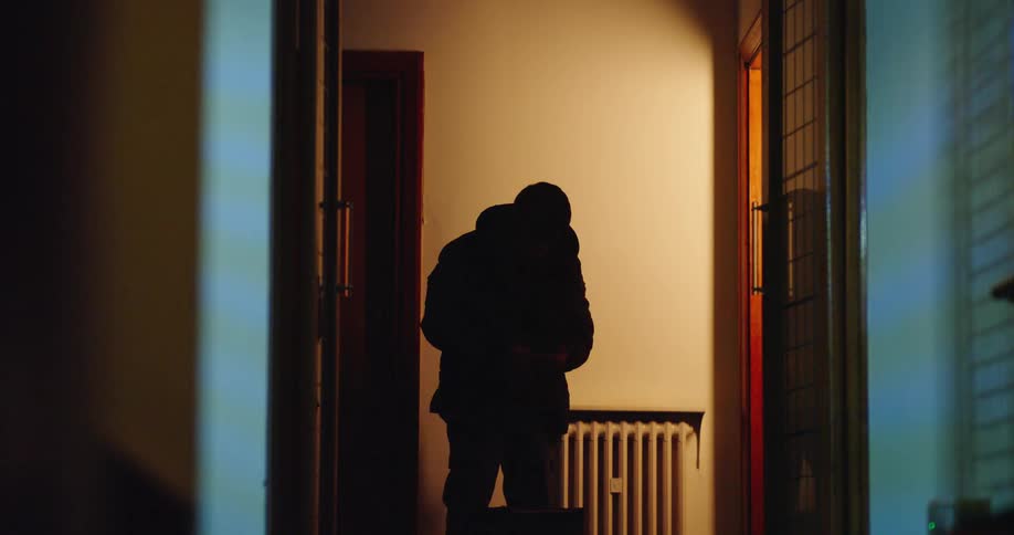 A silhouetted delivery man leaves a paper bag of food on a doorknob in a dimly lit hallway at night