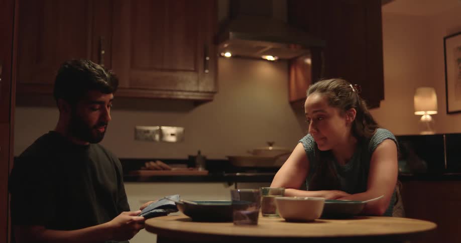 A man unwraps a surprise gift from his partner during dinner to discover a positive pregnancy test, sharing a life-changing moment