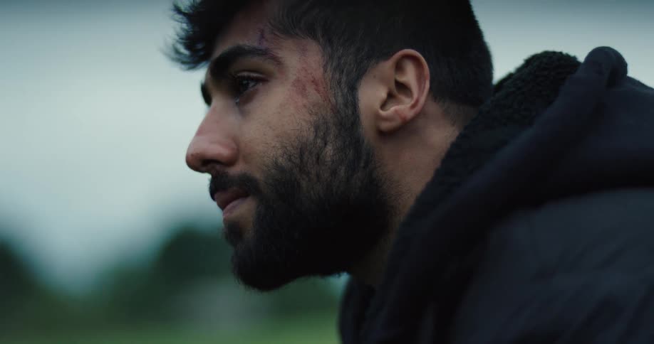 A profile close-up of a young, bearded man with a wound on his face, a tear streaming down as he looks down in sorrow