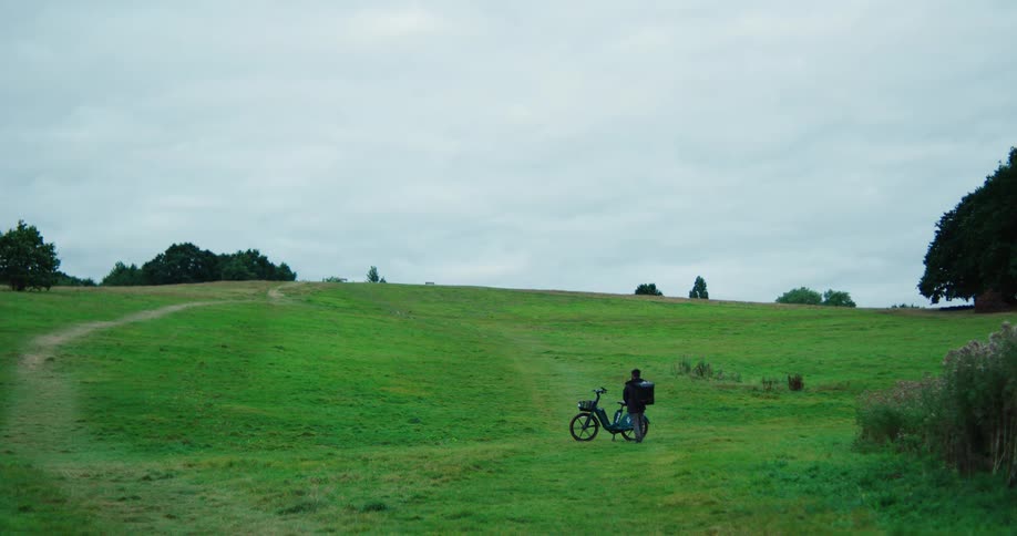 A delivery courier with a backpack pushes his electric bike up a large grassy hill under a cloudy sky