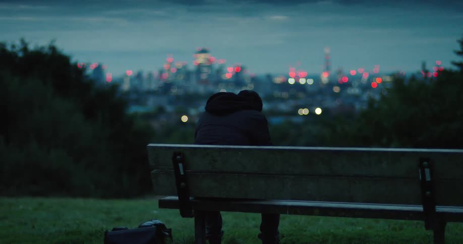 A lonely figure sits on a park bench at dusk, contemplating the distant, illuminated city skyline