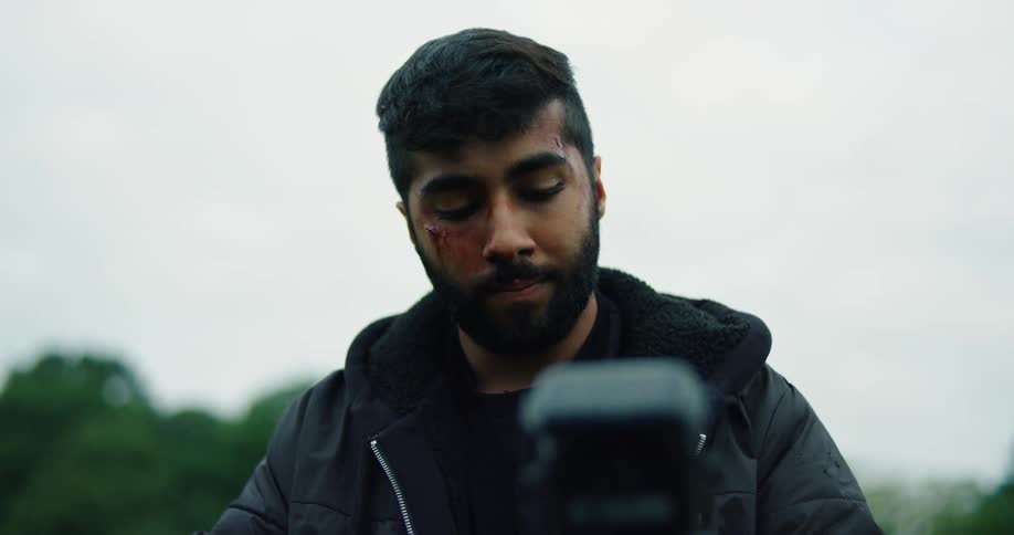 A slow-motion close-up of a young man with a wounded face looking down thoughtfully before gazing up at the overcast sky