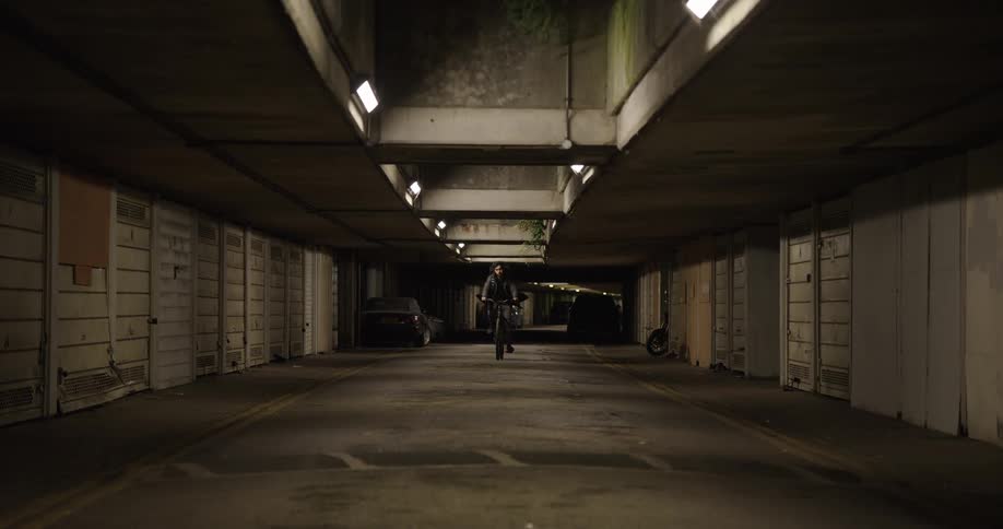 A determined male food courier pedals his bicycle toward the camera through a gritty, dimly lit underground car park at night