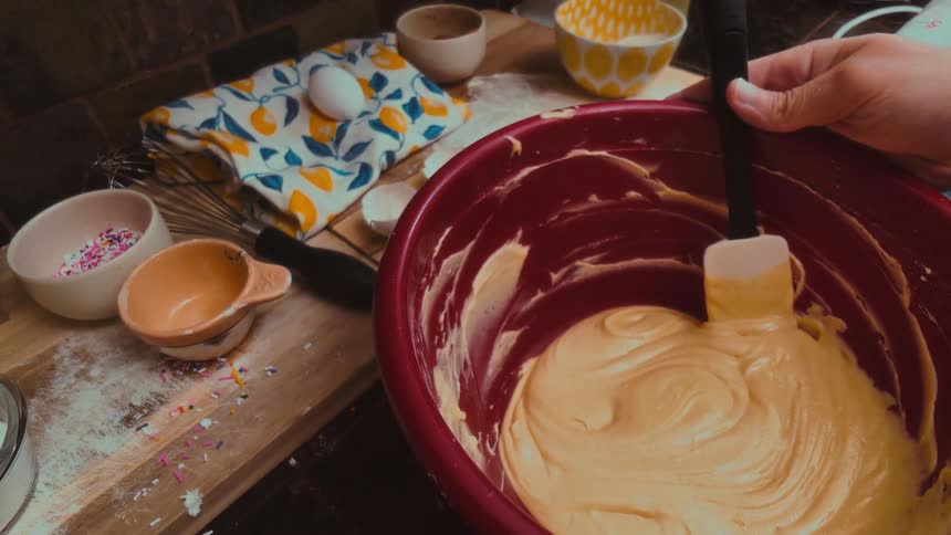 A close-up, slow-motion shot of hands mixing smooth yellow cake batter in a red bowl with a spatula
