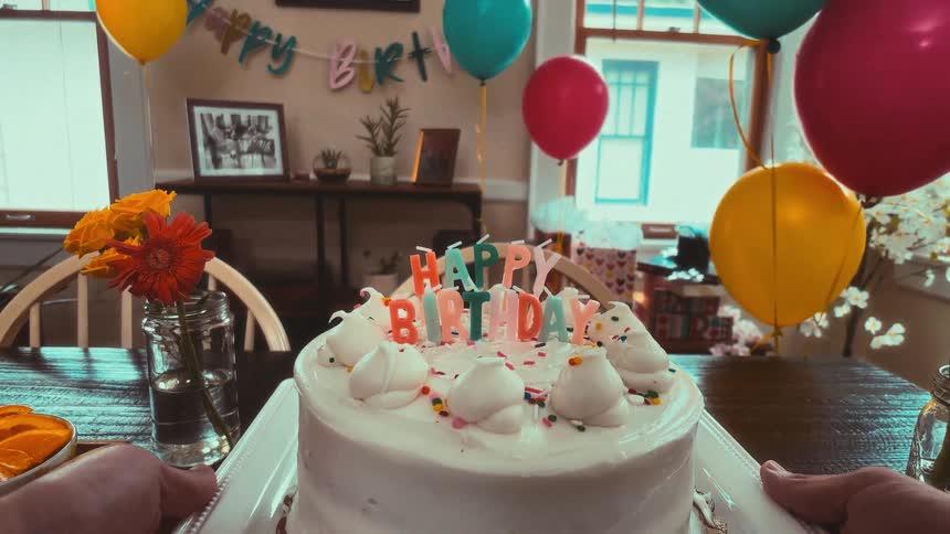 First-person perspective of carrying a birthday cake with 'Happy Birthday' candles and placing it on a dining table in a decorated room