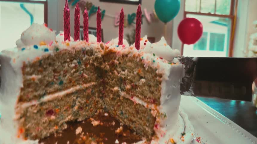 A large kitchen knife slowly slices through a funfetti birthday cake with white frosting and sprinkles, revealing its colorful interior at a party