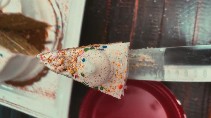 A slow-motion, top-down vertical view of a knife slicing a piece of a festive birthday cake and serving it onto a red plate