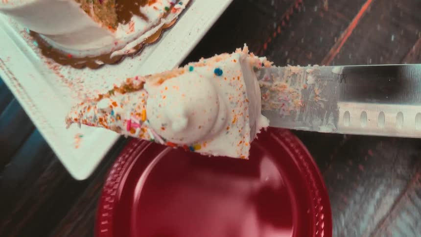 A top-down slow-motion vertical shot of a knife cutting a slice of sprinkle-covered birthday cake and serving it onto a red plate