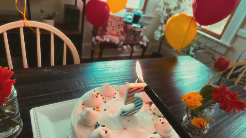 A slow-motion, first-person perspective of a lighter igniting and then extinguishing the number five candle on a festive birthday cake