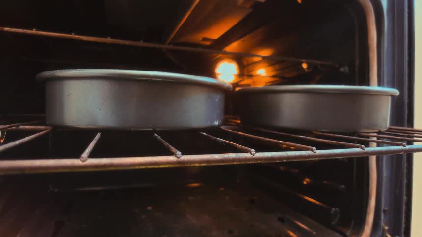 A person wearing oven mitts places two round cake pans into a preheated oven to bake, as seen from an interior perspective