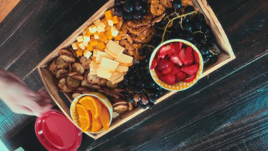 Hands place a vibrant charcuterie board filled with fresh fruits, cheeses, and crackers onto a rustic wooden table, seen from a top-down perspective