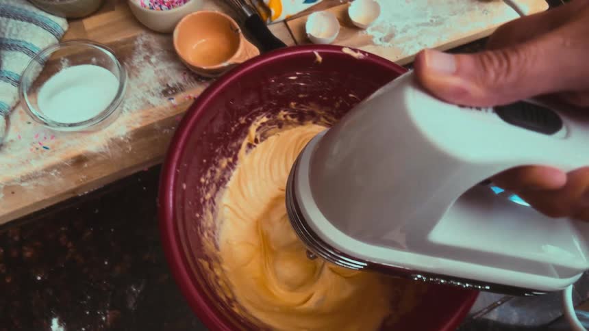 A slow-motion, first-person view of mixing yellow cake batter with a hand mixer in a bowl on a messy kitchen counter