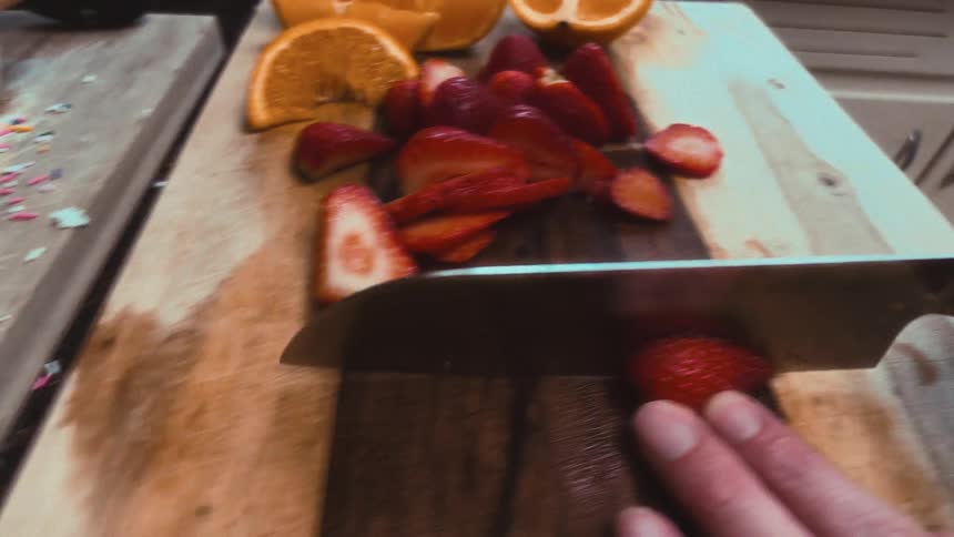 A shaky, first-person perspective captures a hand slicing fresh strawberries on a wooden cutting board next to orange slices