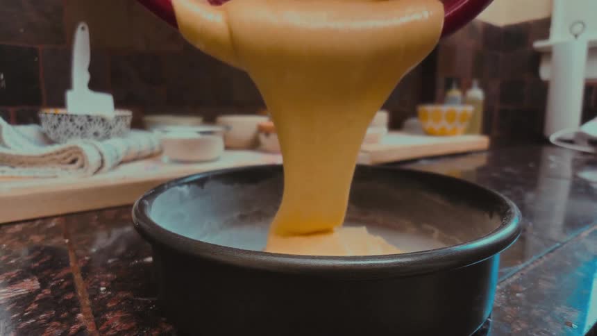 Thick, yellow cake batter is poured from a mixing bowl into a baking pan in a kitchen in slow motion
