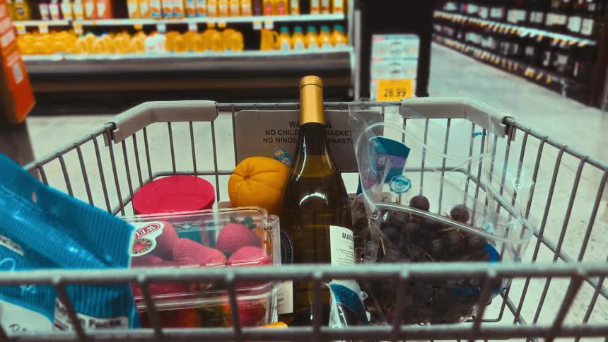 POV of a shopping cart with wine and fresh fruit being pushed through a grocery store aisle