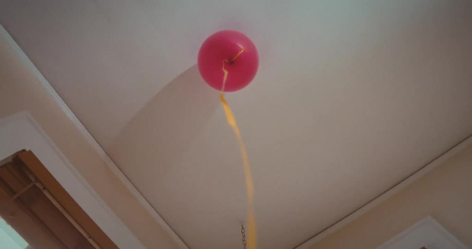 Slow zoom out of a single pink helium balloon with a yellow ribbon floating against the ceiling in a quiet room