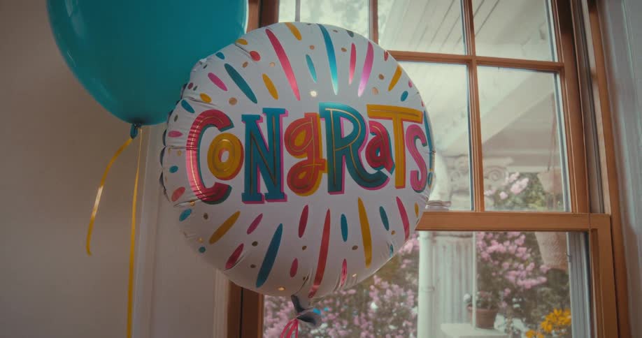 A festive 'Congrats!' balloon floats gently by a window with a bright, sunny porch in the background