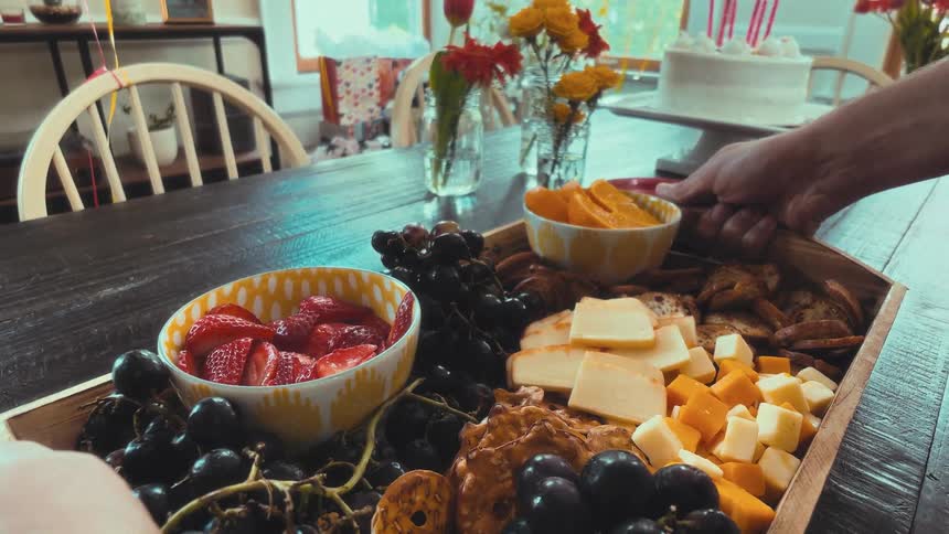 A first-person view of someone carrying a large tray of fruit and cheese to a table set for a birthday party