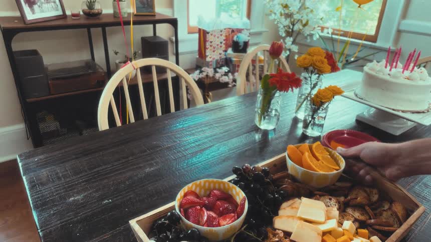 A first-person point of view shot of carrying a charcuterie board to a dining table set for a birthday party with a cake and flowers
