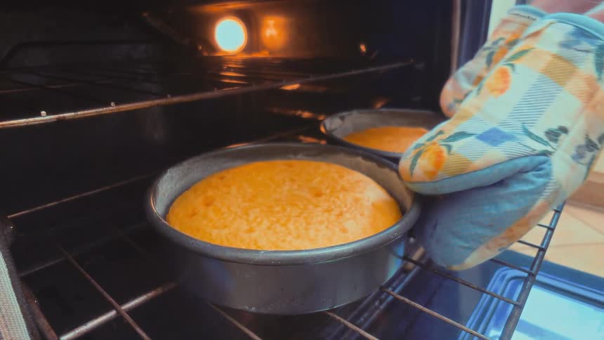 A person wearing an oven mitt pulls a freshly baked cake out of the hot oven, revealing the empty rack
