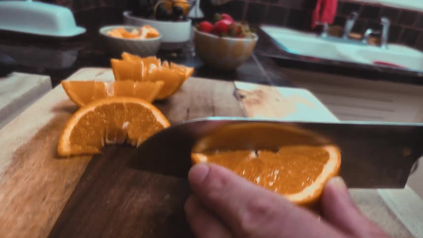 A stylized, shaky POV shot follows a knife blade as it slices a fresh orange on a wooden cutting board in a kitchen