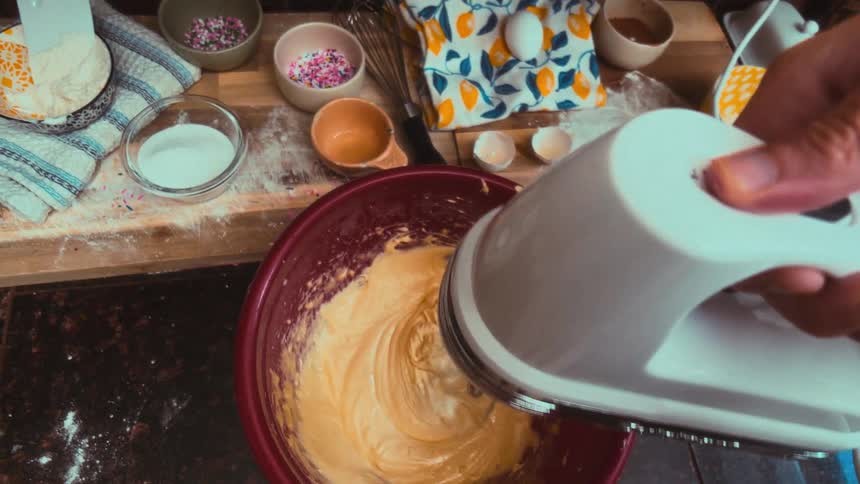 A first-person perspective captures a baker using a hand mixer to blend cake batter in a cozy, messy kitchen
