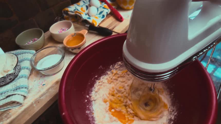 A dynamic top-down shot shows an electric mixer blending flour and an egg into batter in a red bowl for a baking recipe