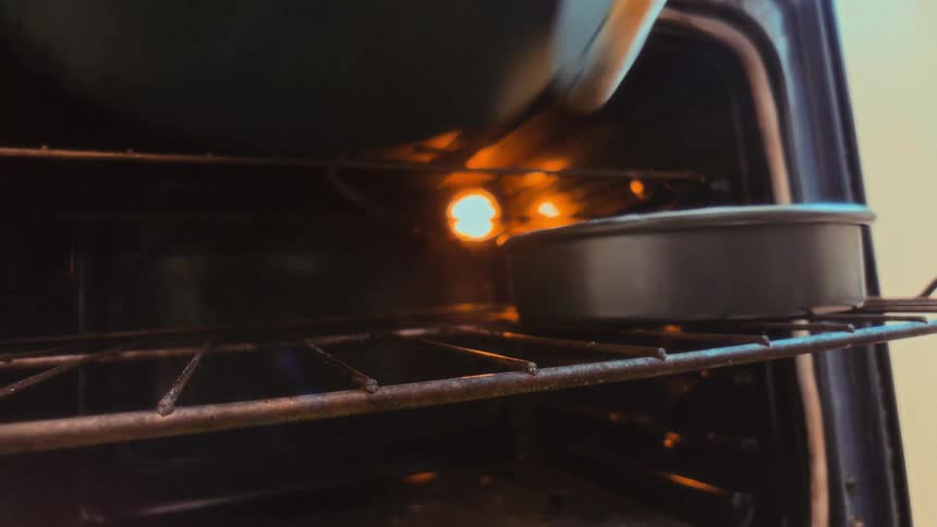 From inside a preheated oven, a person wearing oven mitts places two round baking pans onto the rack to bake