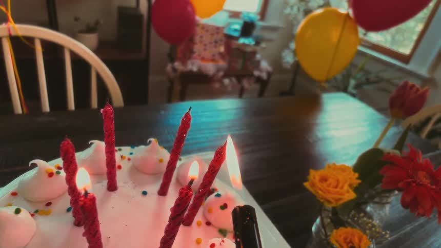 A dreamy slow-motion POV shot of lighting red candles on a white birthday cake surrounded by colorful balloons and flowers