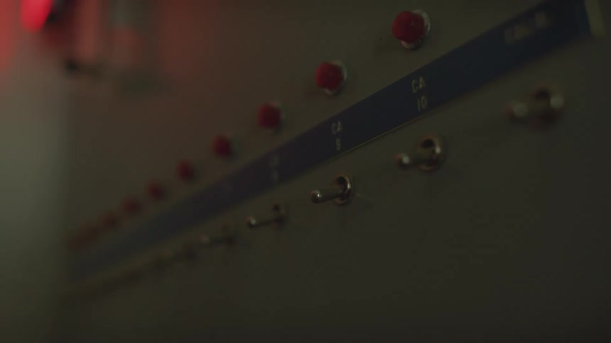A hand flips multiple toggle switches on a control panel as red alarm lights flash in a dark room