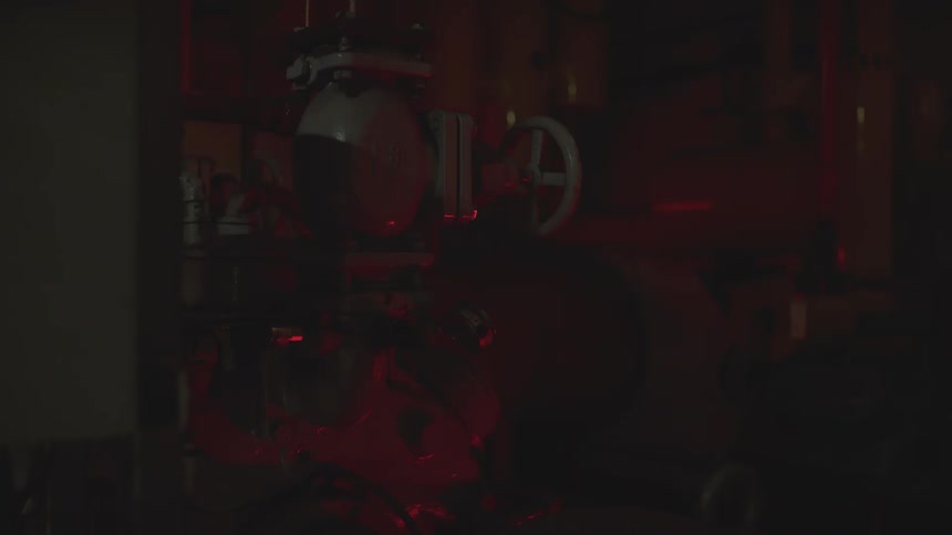 A flashing red alarm light illuminates pipes and valves in a dark industrial engine room