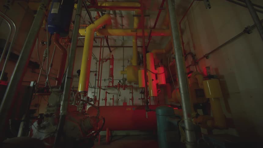 A slow tilt down of an industrial boiler room with complex piping and machinery, illuminated by a flashing red emergency light