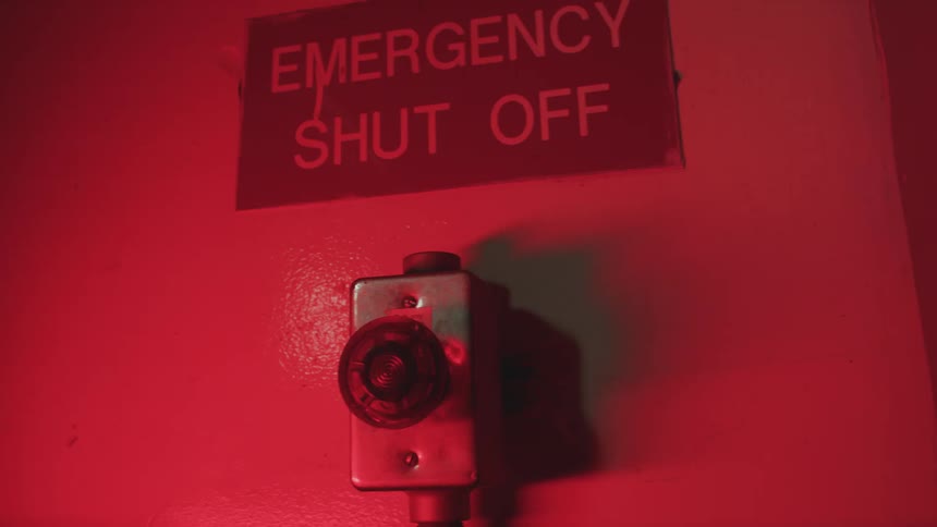 A flashing red warning light illuminates an 'Emergency Shut Off' sign on a wall, signaling an alert
