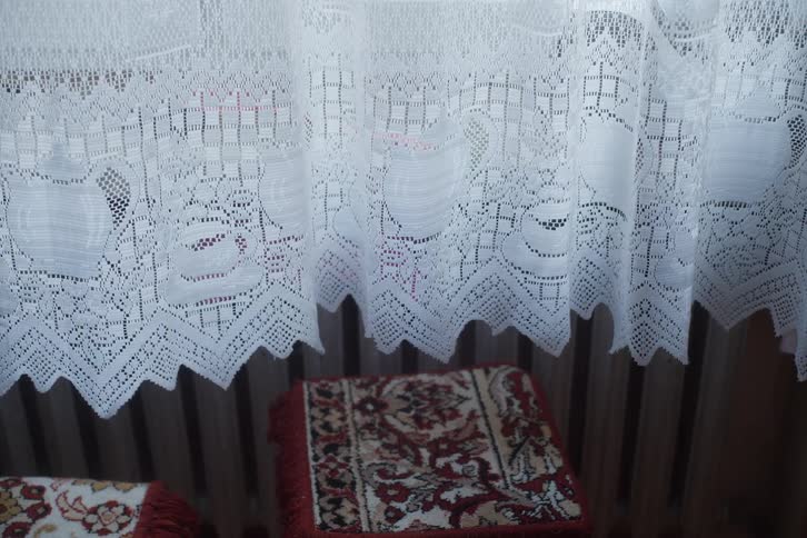 A white lace curtain with a decorative scalloped edge hangs in front of a window and radiator in a cozy room